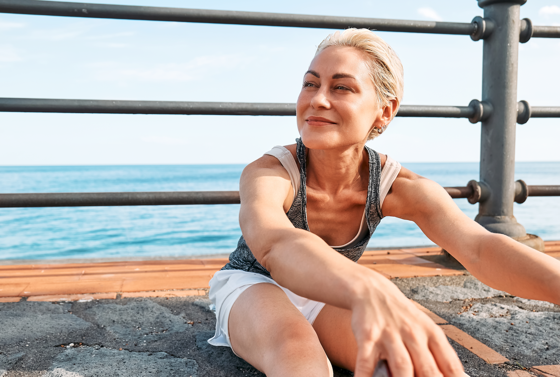 Blonde woman with short hair smiles and stretches by the water after a run.